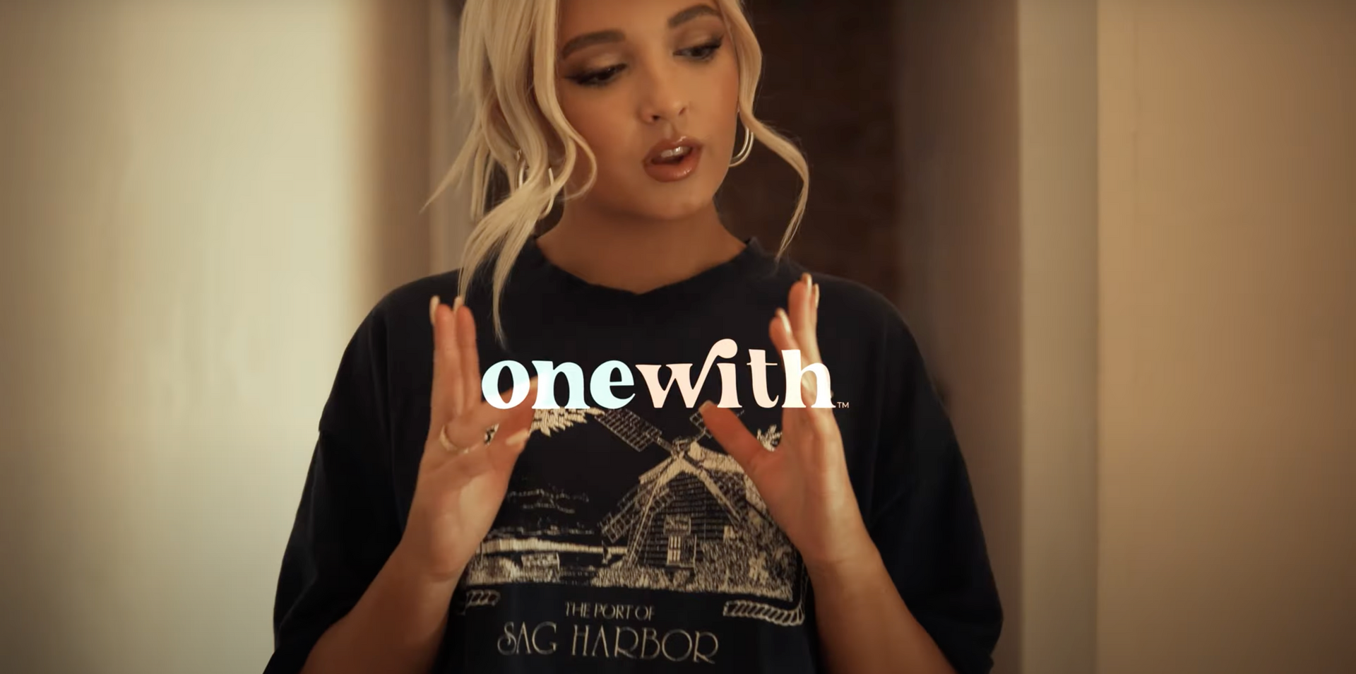 Blonde woman gesturing in a warm hallway wearing an oversized black graphic t-shirt with onewith logo overlaid