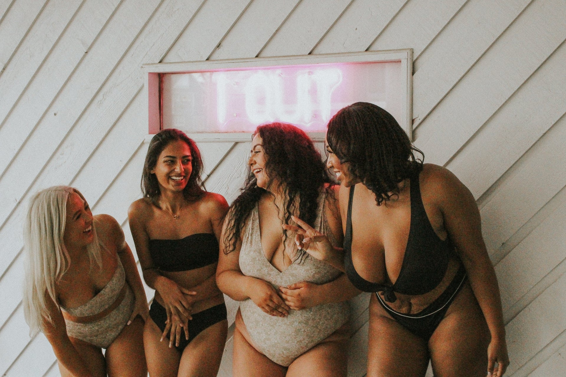 Four women in swimsuits laughing together beside a white diagonal-paneled wall with a pink neon sign.