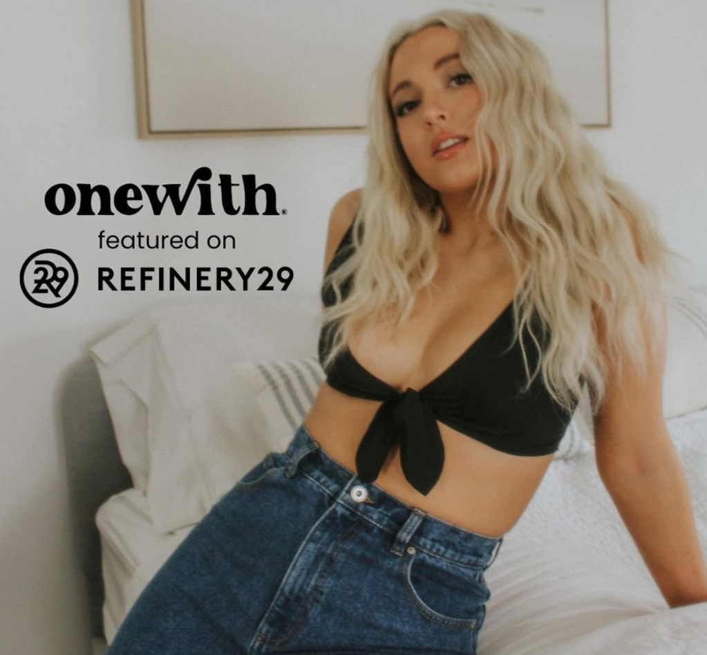 Model reclining on a bed in a black tie-front bikini and high-waisted jeans, overlay text onewith featured on REFINERY29