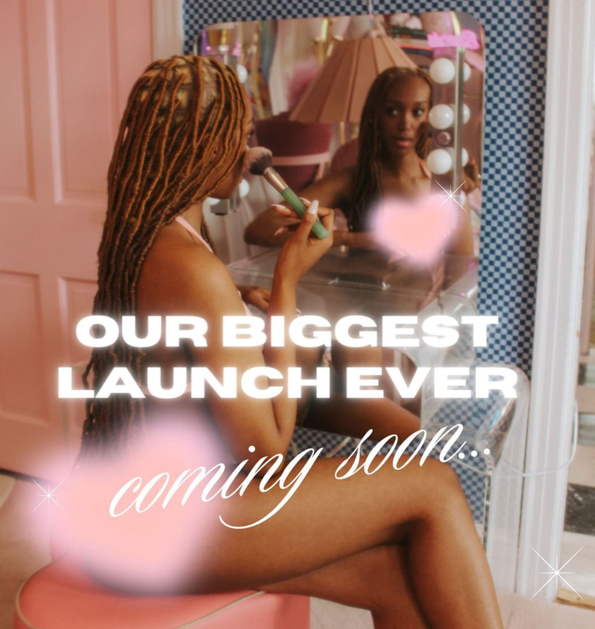 Woman applying makeup at a vanity, reflected in a mirror; text overlay reads 'OUR BIGGEST LAUNCH EVER' and 'coming soon...'