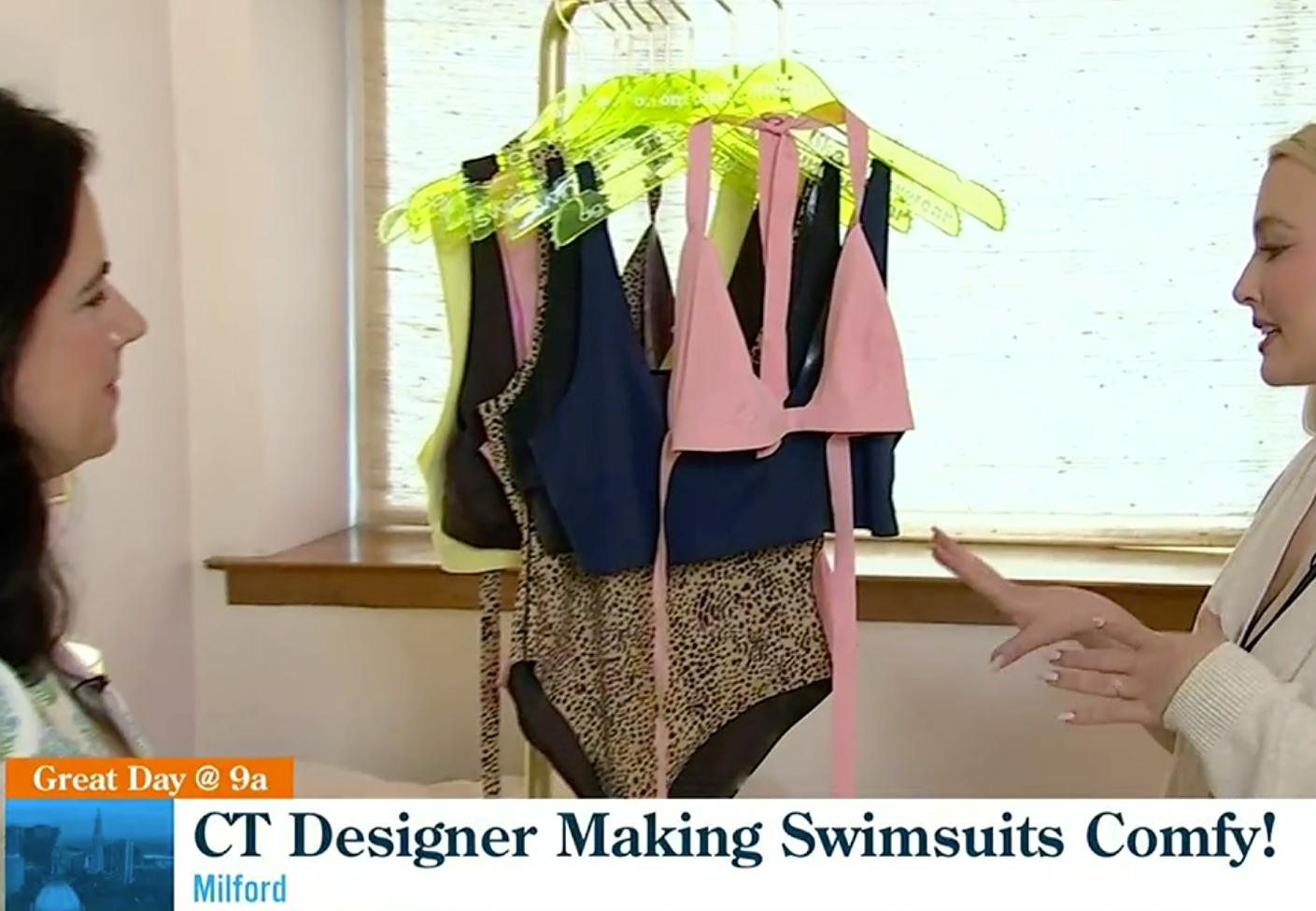 Two women beside a rack of swimsuits on neon hangers; lower-third reads "CT Designer Making Swimsuits Comfy!", "Great Day @ 9a", "Milford".
