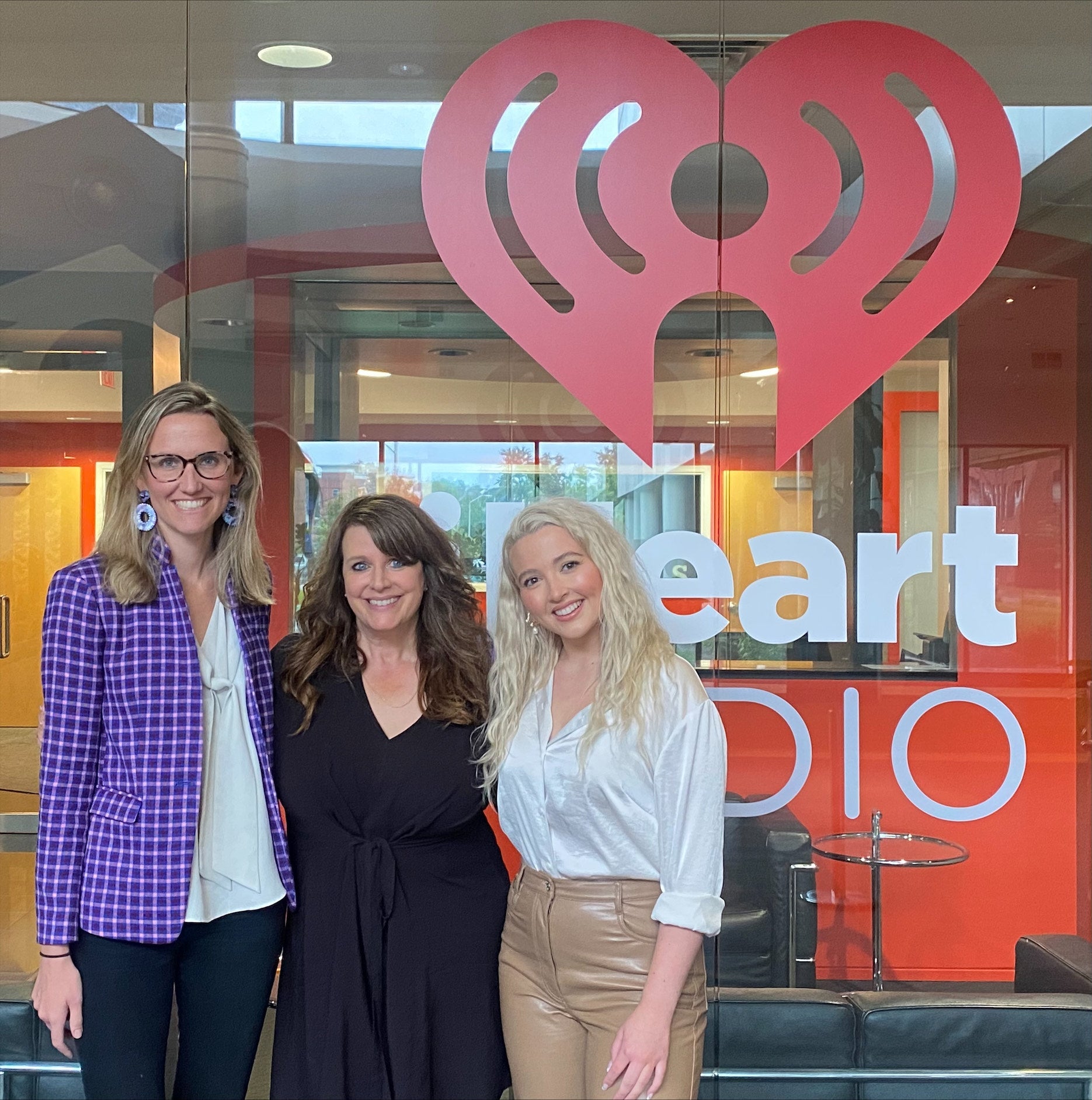 Three women smiling in front of a glass wall with a red iHeartRadio heart logo and the words 'heart radio'.