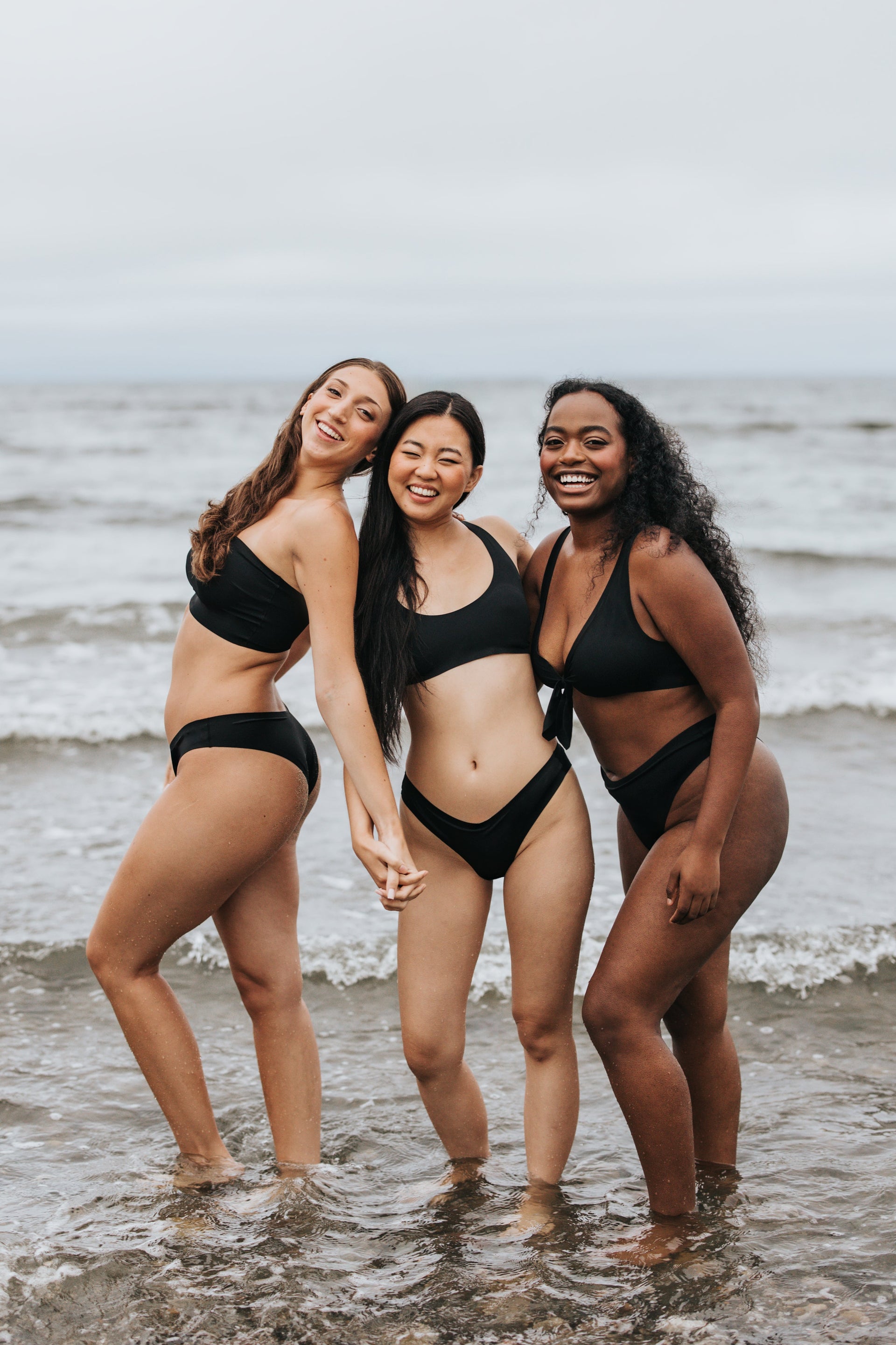 Three smiling women in black bikinis standing hand-in-hand in shallow ocean waves