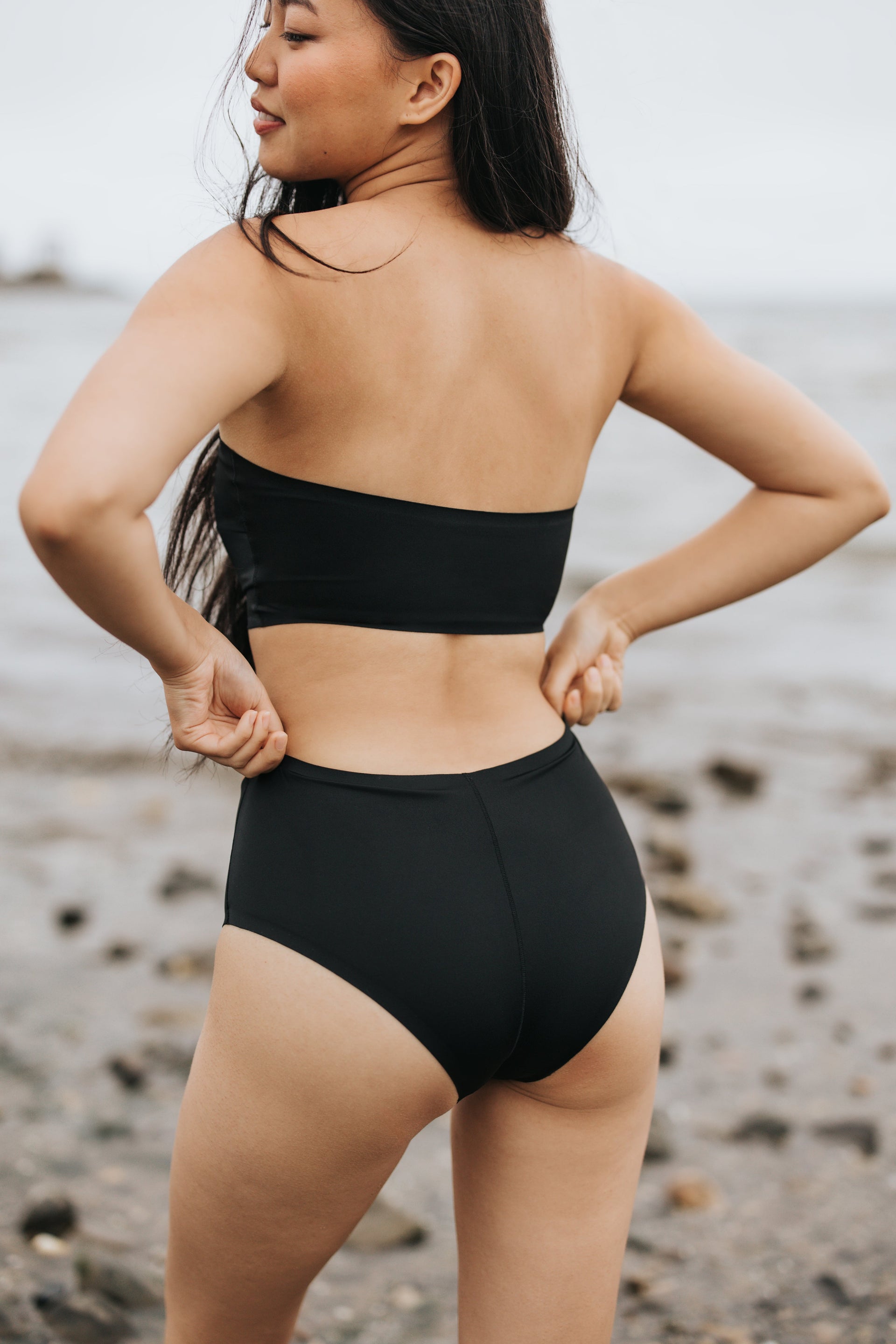 Rear view of a woman adjusting black high‑waist bikini bottoms on a rocky beach
