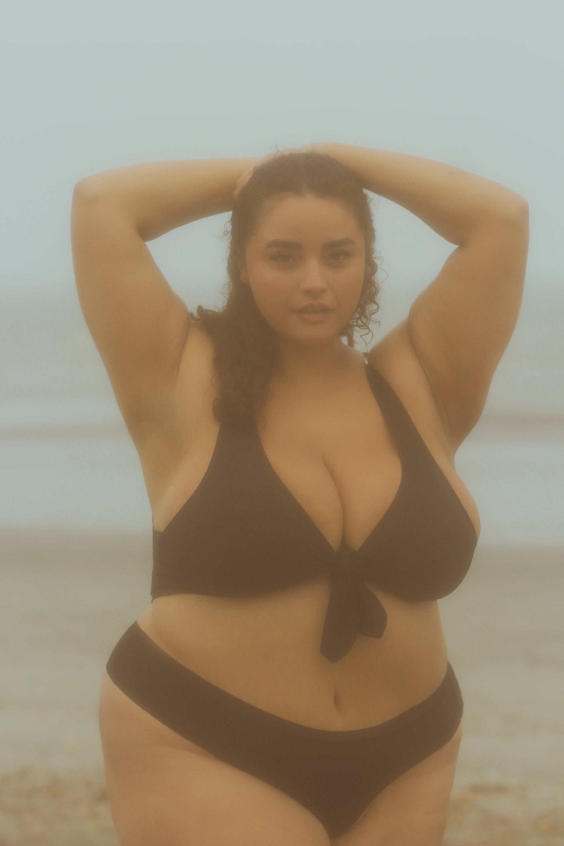 Plus-size woman in a black bikini standing on a foggy beach, hands behind her head, looking at the camera.