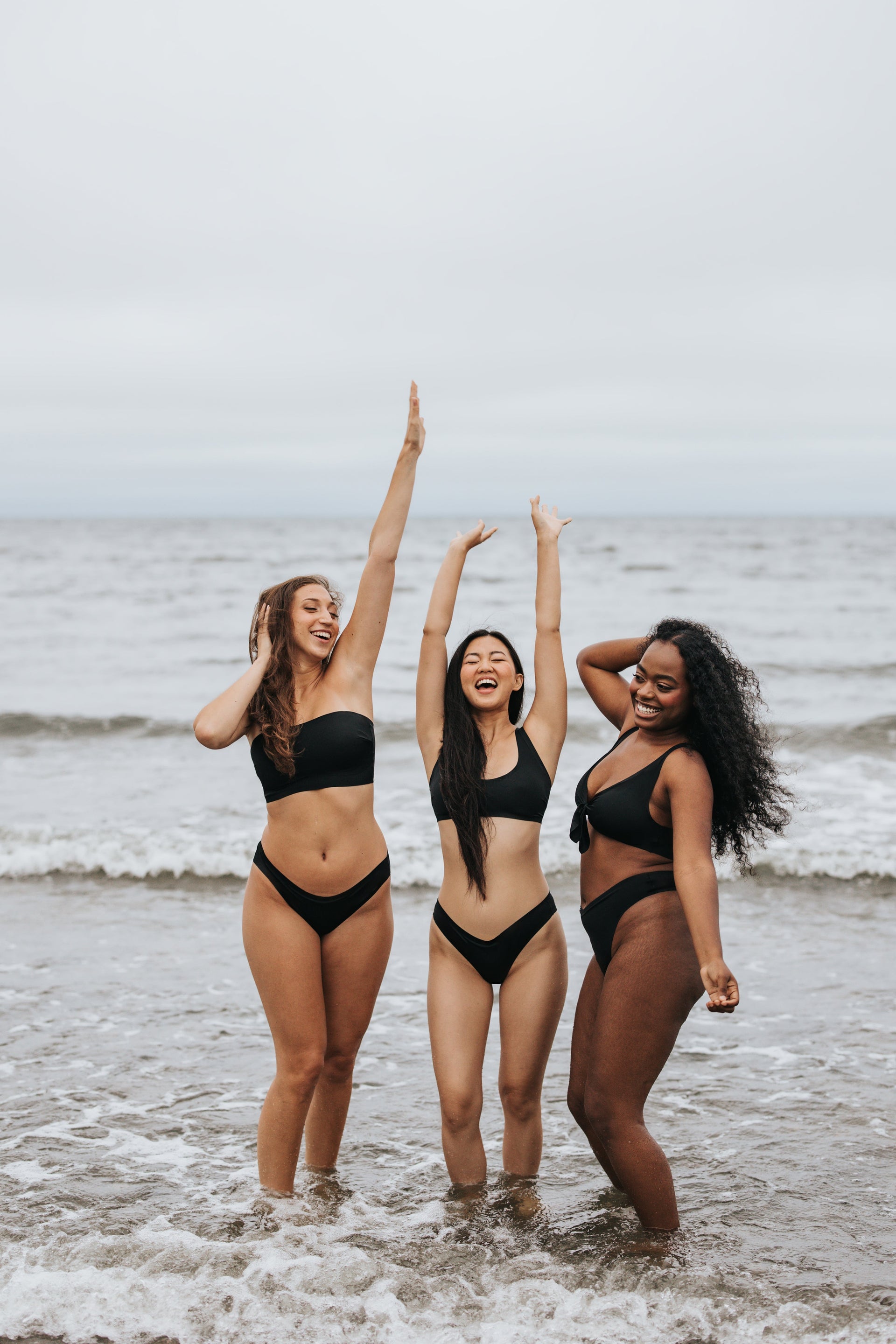Three smiling women in black bikinis standing in ocean surf, arms raised under an overcast sky.