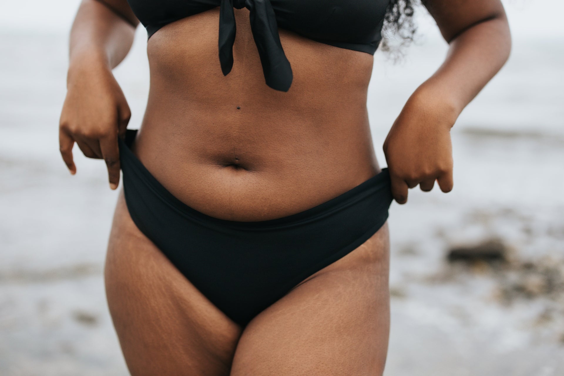 Black woman's torso in black bikini bottoms at the beach, hands adjusting waistband; stretch marks visible.