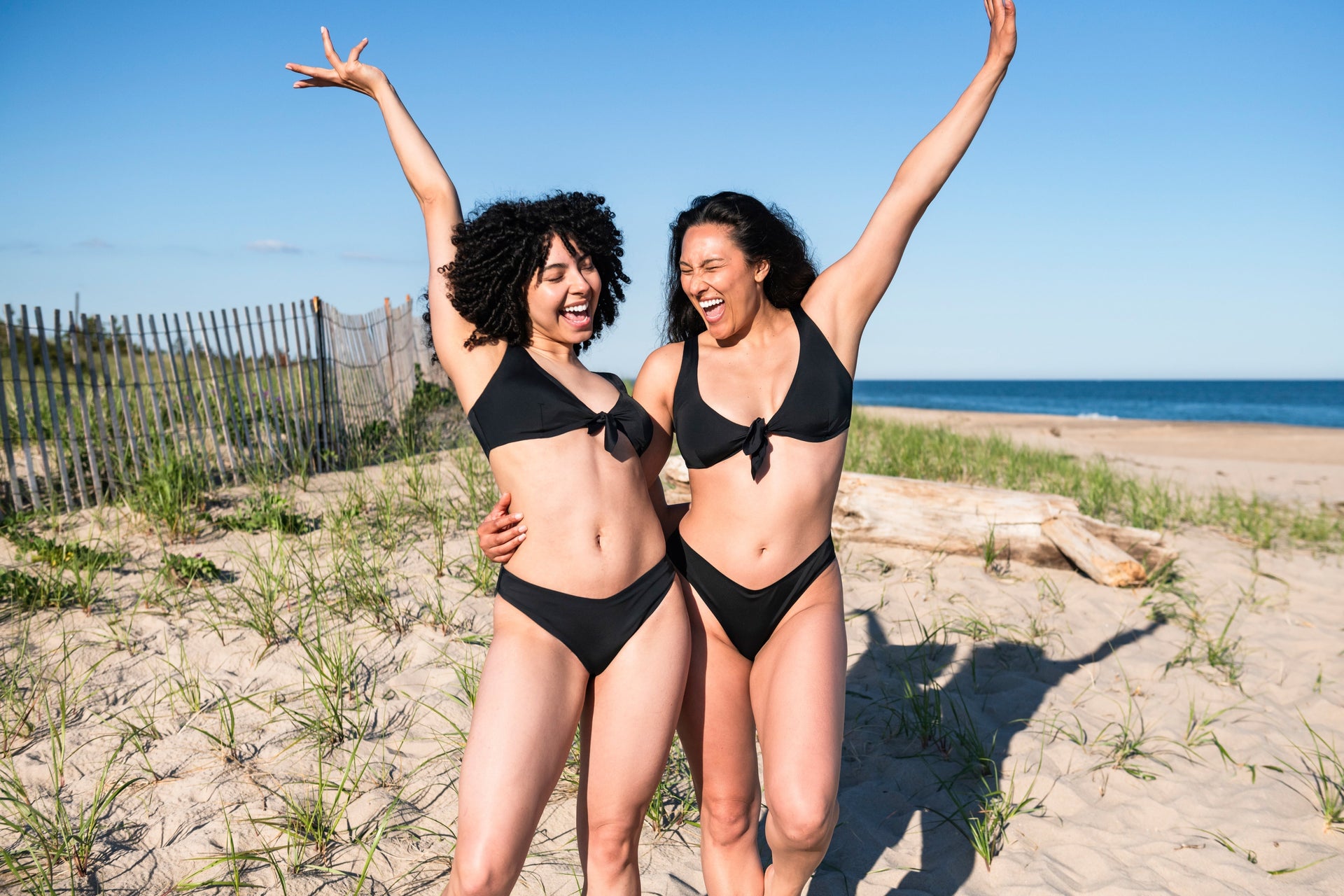 Two women wearing matching black bikinis laugh with arms raised on a sandy beach near a dune fence.