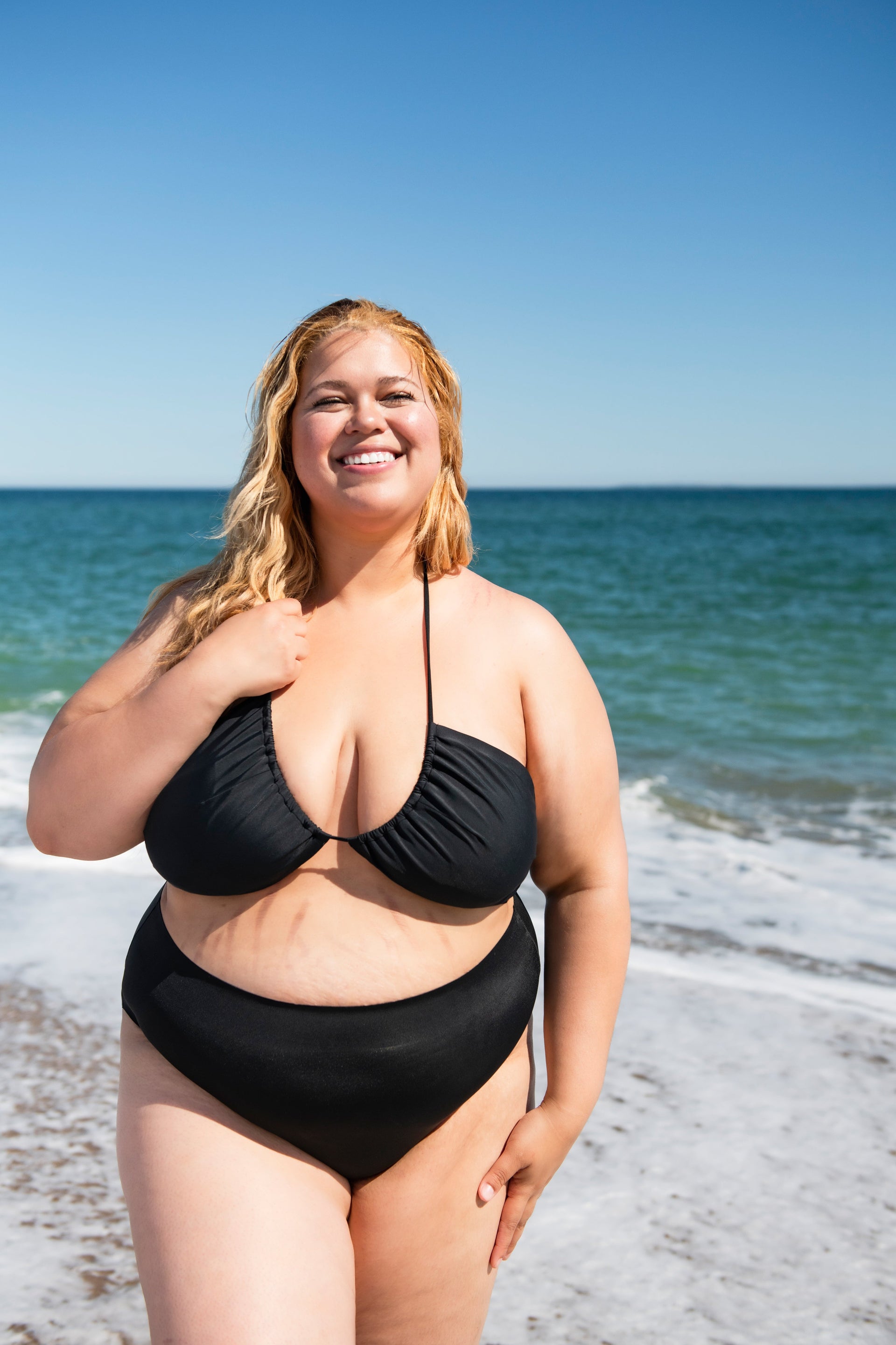 Front view of black tie-front bikini top and high-waisted bottoms on a plus-size model showcasing supportive plunge neckline on a sunny ocean beach
