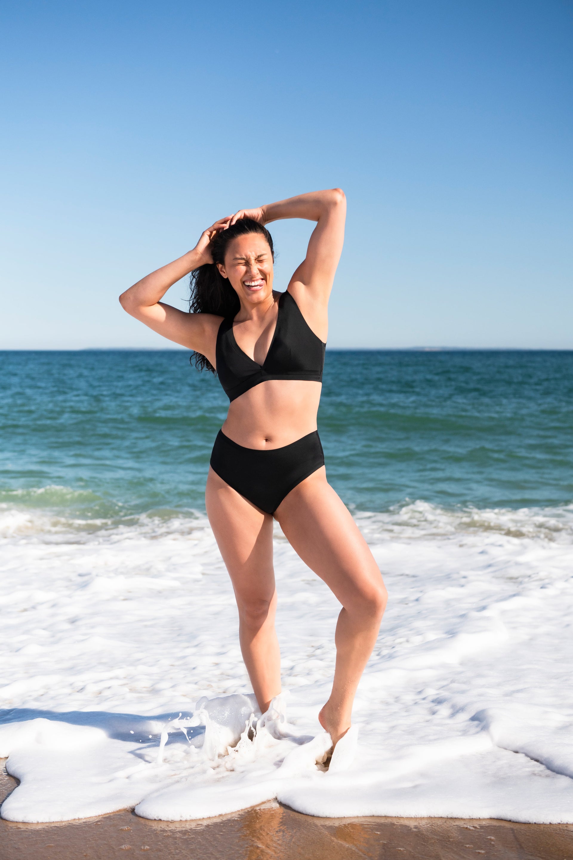 Woman in black two-piece swimsuit laughing with hands in hair as waves wash her feet on a sunny beach