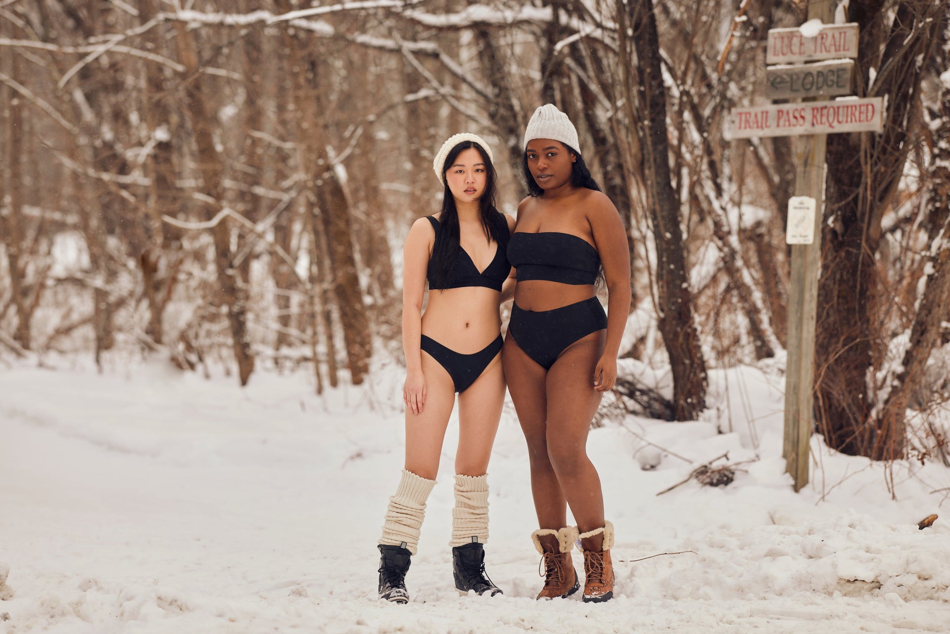 Front of two models wearing black bikinis: V-neck mid-rise and strapless bandeau high-rise in snowy woods
