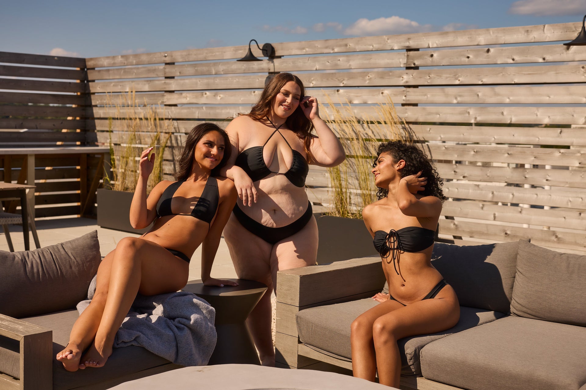 Three women in black bikinis, including a plus-size woman at center, relaxing on a rooftop lounge with wooden fence.