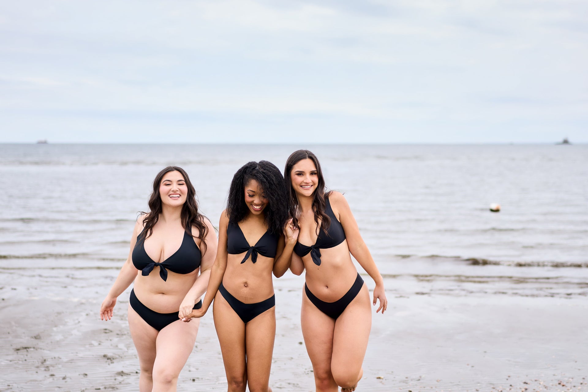 Three women in black bikinis walking hand-in-hand along a calm sandy beach.