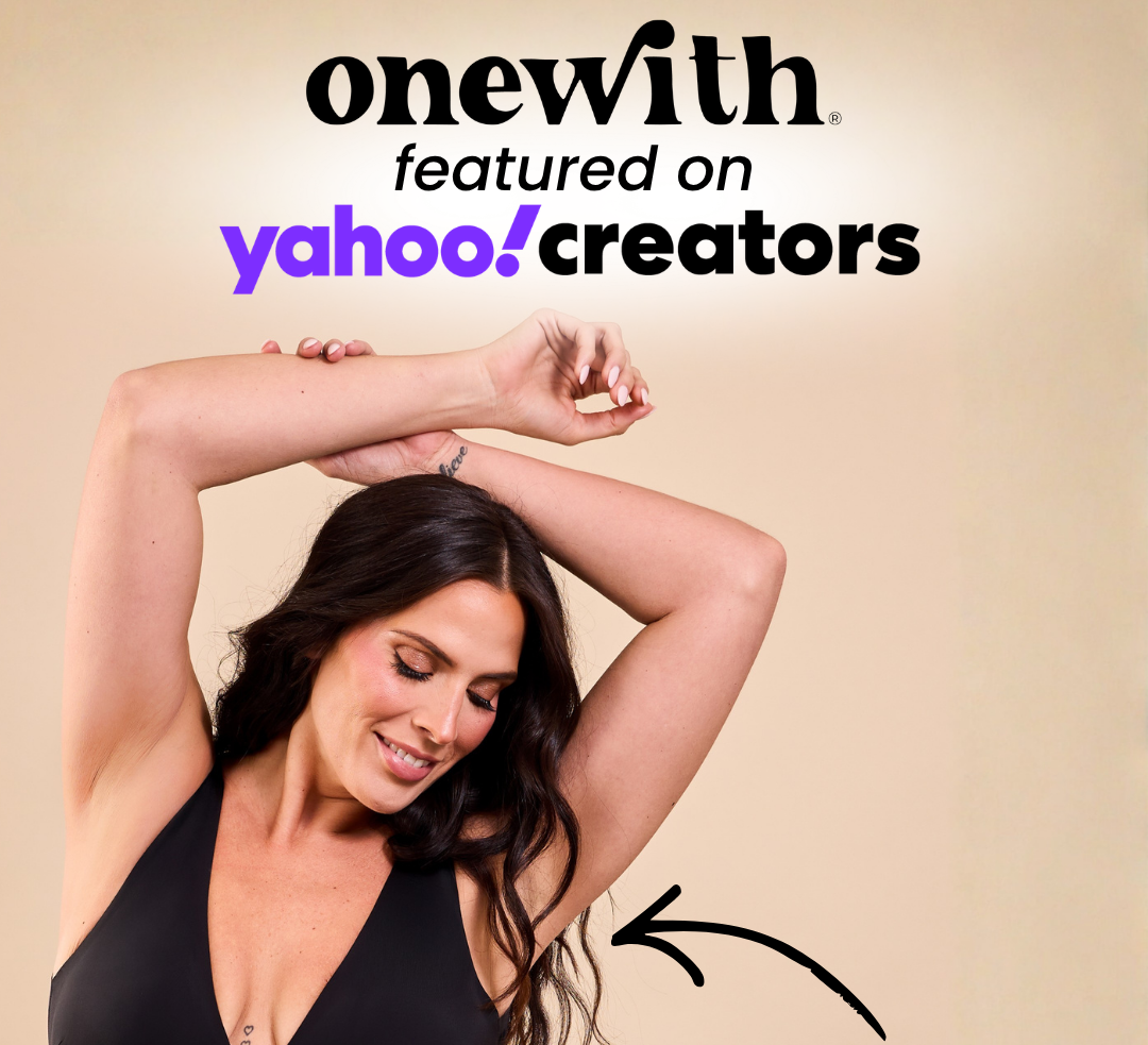 Model in a black plunge one-piece swimsuit raising her arms and smiling against a beige background