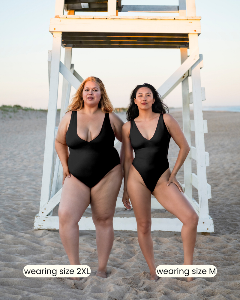 Two women wearing black swimsuits on a beach with a lifeguard stand in the background.