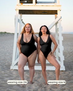 Two women wearing black swimsuits on a beach with a lifeguard stand in the background.