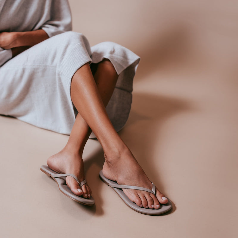 Person seated with legs crossed wearing white top and flip-flops on a beige surface