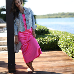 Woman in denim jacket and bright pink skirt sitting on a wooden dock by a lake with trees in the background.