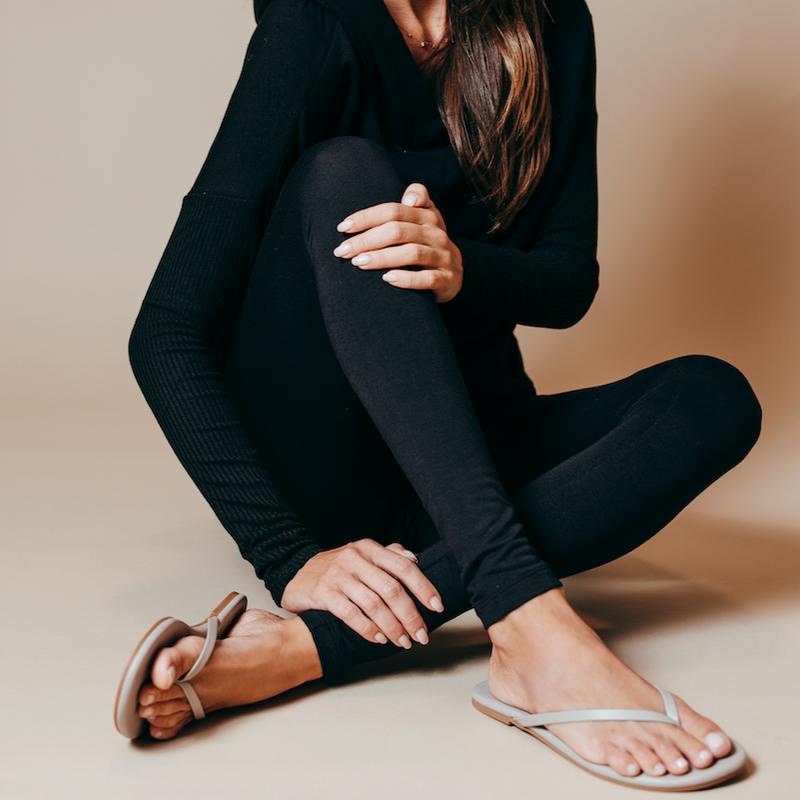 Person seated, adjusting tan flip-flops while wearing a black long-sleeve top and checked trousers.