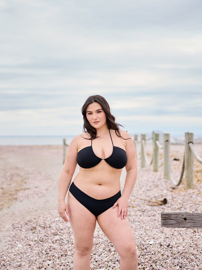 Woman in black bikini standing on sandy beach path near wooden posts under a cloudy sky