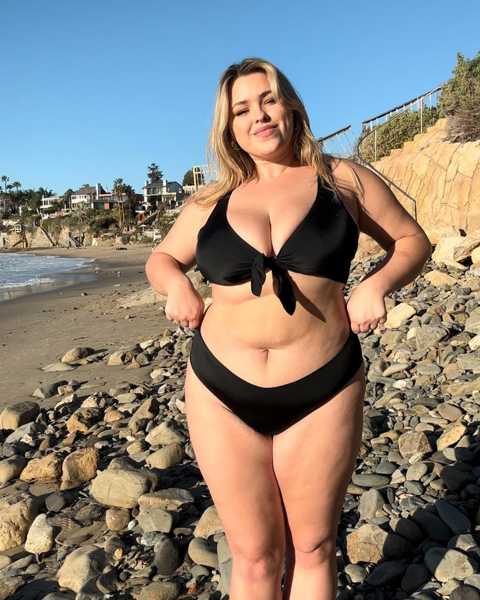 Woman in black bikini standing on rocky beach, smiling; ocean and palm trees in background.
